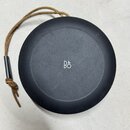 Černý kulatý reproduktor s matným povrchem. Uprostřed je bílé logo B&O. K přístroji je připevněn hnědý šňůrkový řemínek s černým plastovým prvkem, na…