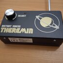 Černý obdélníkový přístroj s nápisem DISTANT VOICES THEREMIN a grafikou planety s prstencem. Na horní straně je stříbrný ovládací knoflík a nápis ADJ…