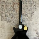 Zadní pohled na černou elektrickou kytaru s tvarem typu Les Paul. Na zadní straně korpusu jsou dva zlaté kryty elektroniky a u krku je vidět kovová s…