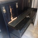 Černý klavír s dřevěným korpusem a ozdobnými zlatými prvky v horní části. Povrch je výrazně opotřebovaný, s velkým odloupaným místem laku na levé str…