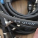 Černý audio kabel s bílým nápisem Cordial GERMANY MICRO. V pozadí jsou vidět další svazky kabelů a konektory, včetně jednoho úhlového jacku. Povrch k…