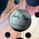 Detailní záběr na kulatý kovový štítek vložený do otvoru v malovaném dřevěném povrchu. Na štítku je černý nápis „Harley Benton“, pod ním text „est. 1…
