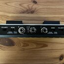 Zadní strana černého elektronického zařízení v kovovém rámu. Na panelu jsou tři jackové konektory označené jako B, A & B a A. Vlevo i vpravo se nachá…