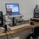 Na dřevěném stole je umístěno studio s notebookem na stojanu, dvěma černými studiovými monitory s nápisem M-Audio a bílou klávesnicí. V popředí leží…
