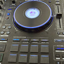 Černý DJ mixér s nápisem PRIME 4+. V horní části jsou umístěny kontrolní knoflíky a přepínače, ve středu se nachází velký jog-wheel s modrým podsvíce…