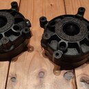 Dvě černé kulaté součástky, pravděpodobně reproduktory nebo snímače, s plastovým nebo kovovým rámem s výstupky pro šroubování. Uprostřed každého kusu…
