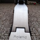 Fotografie zachycuje pedál bicích s kovovou stříbrnou ploščkou a černým základním mechanismem. Na spodní části ploščky je vyražen nápis Roland. Povrc…