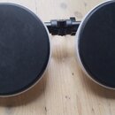 Dvě černé kulaté plochy s bílým okrajem, které jsou spojeny černou kovovou tyčí s upínacím mechanismem. Celý předmět leží na světlém dřevěném povrchu…