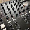 Černý DJ mixér s kovovým povrchem a řadou ovládacích prvků. Jsou vidět posuvníky, otočné knoflíky a tlačítka s nápisy GAIN, FILTER, CUE, AUX, CUE MIX…