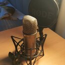 Kondenzátorový mikrofon stříbrné barvy v černém pavoukovém držáku. Za mikrofonem je umístěn černý popfilter s nápisem RØDE®. Povrch mikrofonu a držák…