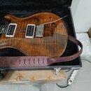 Elektrická kytara s hnědým plamenovitým topem a dvěma humbuckery v bílých rámečcích leží v černém tvrdém kufru. Na hmatníku jsou vidět vkládače ve tv…