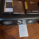 Černý studiový monitor s dvěma basovými reproduktory a jedním výškem vprostřed, který nese logo ADAM. Na horní části přístroje leží tři černé dokumen…