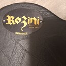 Černý kufřík z texturovaného materiálu s oválným logem. Na logu je zlatý nápis „Rozini“ a pod ním menší text „DESDE 1995“. Pod oválem je přímo na kuf…