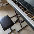 Černý digitální piano s klávesnicí na černém X-stojanu značky Soundbeam. K nástroji patří černá polstrovaná stolečka s nápisem CASIO na jedné z nožek…