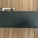 Černý obdélníkový přístroj s kovovými úchyty po stranách. Uprostřed je viditelné logo v podobě trojúhelníku a pod ním nápis behringer. Povrch je matn…