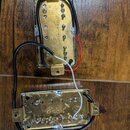 Dvě elektrické kytarové snímače v kovových krytech zlaté barvy. Na obou je vyryt nápis Belcat a logo ve tvaru písmene B v kruhu. Povrch krytů vykazuj…