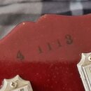 Detail červeného povrchu hlavy hudebního nástroje. Na laku je vyraženo sériové číslo 4 1113. V dolní části jsou vidět části dvou kovových ladíček s v…