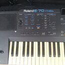 Šedý elektronický klavír s nápisem Roland E-70 INTELLIGENT SYNTHESIZER. Přístroj má ovládací panel s množstvím tlačítek, posuvníkem hlasitosti a malý…