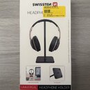 Fotografie znázorňuje bílé papírové balení produktu. Na krabici je nápis SWISSTEN, logo ve tvaru stylizovaného písmene N a text HEADPHONE STAND. Dále…