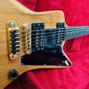Detail elektrické kytary s přírodním dřevěným povrchem a černým pickguardem. Nástroj má dva humbuckery, zlatý most s hrazdiky a dvě černé ovládací kn…