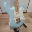 Světle modrá elektrická kytara typu Stratocaster s bílým pickguardem a hnedým hmatníkem. Nástroj má tři snímače typu single-coil, jeden ovládací knof…