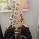Fotografie zachycuje hlavu elektrické kytary z přírodního světlého dřeva s chromovanými ladičkami. Na hlavě je černý nápis Squier MINI by Fender. Kyt…