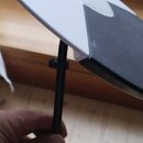 Fotografie zachycuje černý kovový stojan s úchytem, který drží bílý kulatý disk s černým prstencem a středovým kolíkem. Disk má v pravé části černý p…