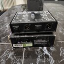 Černý box s ovládacími prvky a konektory, na kterém je bílým nápisem PDB. Na přední straně jsou dva přepínače a dva XLR konektory. Zařízení leží na k…