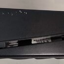 Černý plastový stojan na noty v zavřeném stavu. Povrch vykazuje drobné oděrky a světlé škrábance v horní části. Na předmětu nejsou viditelné žádné zn…