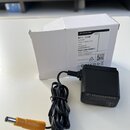 Černý síťový adaptér značky SENNHEISER s nápisem NT 2-3 CW Switching Power Supply a žlutým konektorem. Vedle adaptéru je bílá papírová krabice s text…
