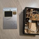 Fotografie ukazuje otevřený černý přístroj s viditelnými vnitřními elektronickými součástmi, zapojením a dráty na desce plošných spojů. Vedle přístro…