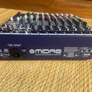 Modrý mixážní pult s řadou posuvníků, knoflíků a konektorů na horní straně. Na zadní straně je nápis MIDAS a text DESIGNED AND ENGINEERED IN ENGLAND.…
