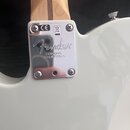 Detail krku a těla bílé elektrické kytary s kovovým krytem spojení. Na chromovaném krytu je vyryto logo Fender a nápis CORONA, CALIFORNIA. V horní čá…
