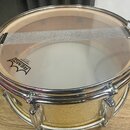 Fotografie znázorňuje snare drum s béžovým, texturovaným povrchem korpusu a chromovaným obručováním. Na blaně je viditelné logo REMO s nápisy AMBASSA…