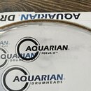 Fotografie zachycuje bílý bubenový kožen s nápisy. Je na něm vidět logo a text AQUARIAN® FOCUS-X ™ oraz opakovaně nápis AQUARIAN® DRUMHEADS. Povrch j…