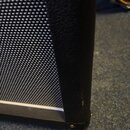 Detailní pohled na roh černého kytarového komba. Je vidět černá pletená textura reproduktorové síťky a hrubý povrch skájového potahu. V dolní části r…