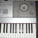 Stříbrný digitální klavír s bílými a černými klávesami. Na ovládacím panelu je nápis Portable Grand DCX-640 a sekce MUSIC DATABASE s seznamem stylů.…