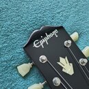 Detail hlavy černé kytary s bílým nápisem Epiphone. Na povrchu je umístěn světlý ornament ve tvaru šipky. Jsou vidět kovové ladičky s krémovými knofl…