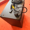 Černý kombozesilovač s povrchem z texturovaného vinylu. Na horní ploše nástroje leží černý spirálový kabel a černý napájecí kabel s žlutým štítkem. N…