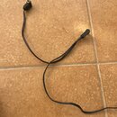 Černý napájecí kabel s konektory na obou koncích. Na jedné straně je kulatý DC konektor, na druhé straně je černý plastový kryt. Na kabelu nejsou vid…