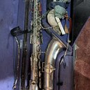 Stříbrný saxofon v otevřeném černém kufru s fialovou výplní. V kufru jsou rovněž šedý hadr, bílý předmět a hnědý popruh. Na nástroji nejsou žádné čit…