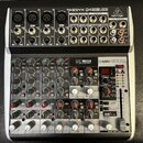 Šedý mixážní pult značky behringer s nápisem XENYX QX1202USB. Zařízení obsahuje několik vstupních kanálů s ovládacími prvky pro GAIN, EQ a FX, USB IN…
