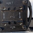 Fotografie zachycuje spodní část černého elektronického zařízení s úchyty po stranách, umístěného v transportním kufru. Na černém povrchu jsou ručně…