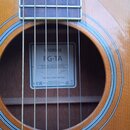 Detail akustické kytary s viditelným hradlem a otvorem rezonátoru. Uvnitř otvoru je bílá štítková nálepka s nápisy YAMAHA, FG-TA a TransAcoustic. Dol…