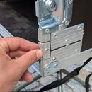 Detailní pohled na roh černého transportního kufru s hliníkovými hranami a kovým zapínáním. Ruka ukazuje na hliníkový profil, který je připevněn šrou…