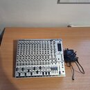 Stříbrný mixážní pult s černými posuvníky a ovládacími prvky. V horní části je uveden nápis BEHRINGER a model EURORACK MX 2642A. Vedle přístroje leží…