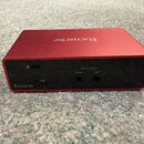 Červený kovový audio interface s nápisem Focusrite na horní straně. Zadní strana je černá a obsahuje USB port, dva výstupy označené LINE OUTPUTS RIGH…