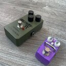 Na fotografii jsou dva efektové pedály na dřevěné podlaze. Větší pedál je olivově zelený s nápisy BIG MUFF π, electro-harmonix a ОВЕРДРАЙВ. Menší ped…