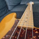 Elektrická basová kytara s přírodním dřevěným povrchem těla a hmatníkem. Nástroj má tmavě červený plexiglasový pickguard s mramorovaným vzorem. Na fo…
