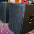 Dva černé reproduktory s kovovými mřížkami a bočními úchopovými rukojetmi. Na spodní části mřížek je umístěno logo JBL. V pozadí je vidět část zaříze…